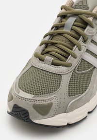 Zapatilla atlética verde oliva con materiales de malla y ante, acentos grises, suela texturizada y tres rayas blancas en el costado.