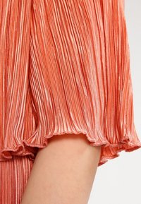 Robe en tissu plissé de couleur corail, avec une texture douce. L'ourlet présente un détail volan élastique et des lignes verticales sur l'ensemble.