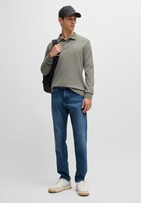 Green long-sleeve polo, denim jeans, white sneakers with beige accents, and a black cap. Model holds a black backpack.
