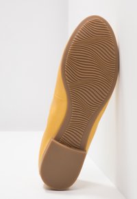 Chaussure jaune avec une tige lisse et une semelle en caoutchouc marron texturée présentant un motif ondulé, posée sur un fond blanc.