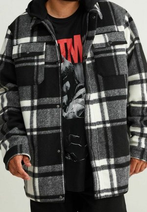 Homme portant une veste à carreaux noire et blanche sur un t-shirt graphique noir avec un texte rouge et un dessin de casque argenté.