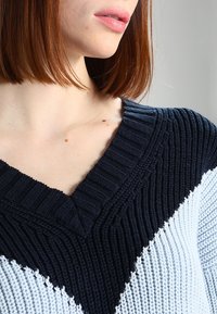 Svetr s výstřihem do V, který se vyznačuje pleteným designem v námořnické modré a světle modré barvě, texturovaným žebrovaným vzorem a chevronovým vzorem na těle.