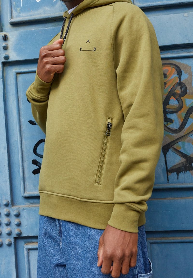 Homme portant un sweat à capuche vert olive avec poche zippée noire et petit logo, debout devant une porte métallique bleue couverte de graffitis.