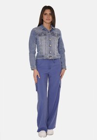 Giacca di jeans con tasche frontali, colore blu chiaro; abbinata a pantaloni cargo larghi di colore lavanda e sneakers bianche.