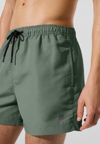 Zelené plavkové šortky s elastickým pásom a sťahovacou šnúrkou, s malým logom na ľavej nohe, vyrobené z ľahkej látky.