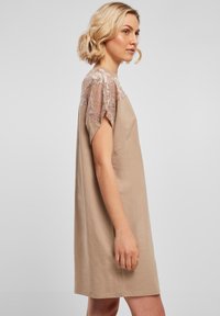 Lühike beež kleit, mis on valmistatud pehmest kangast, lühikeste varrukatega ja pitsdetailidega õlgadel. Lihtne, lahtine lõige.