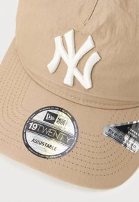 Tan spalvos beisbolo kepuraitė su balta išsiuvinėta 'NY' logo, reguliuojama sagtis, išlenktas vizorius ir etiketės, nurodančios stilių ir prekių ženklą.