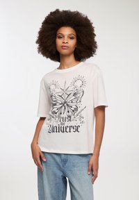 Koszulka w kolorze kremowym z dużym czarnym graficznym motylem, słońcami i napisem "ufaj wszechświatowi". Krótkie rę sleeves, luźny krój.