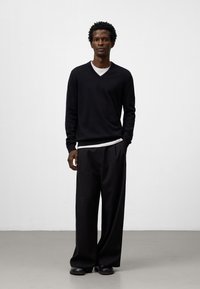 Maglione nero a scollo a V con maniche lunghe, indossato sopra una camicia bianca, abbinato a pantaloni neri larghi e scarpe nere. Sfondo semplice.