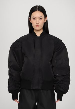 Black bomber jacket with oversized sleeves, ribbed cuffs and hem, front zip closure, and a side zip pocket in a matte, smooth texture.