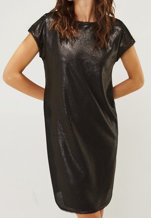 Kvinde iført en kortærmet, knælængde sort palietkjole med rund halsudskæring, stående med hænderne bag ryggen.