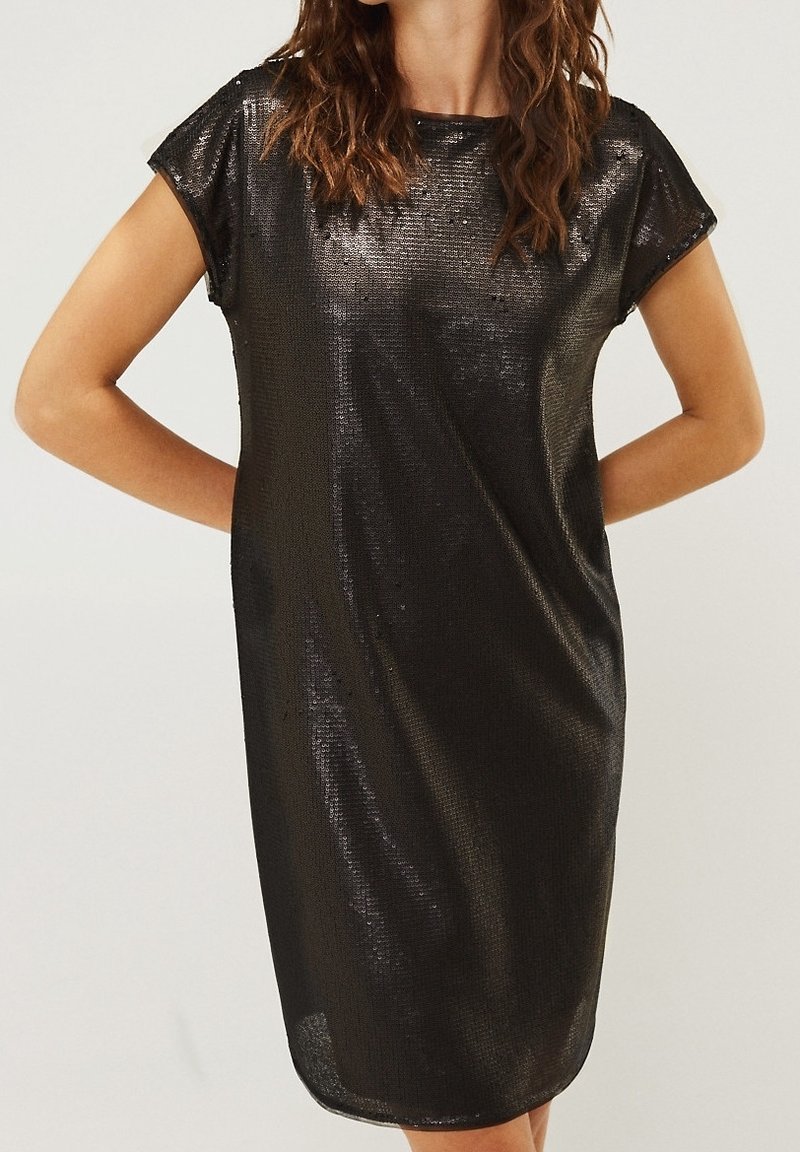 Femme portant une robe noire à paillettes, manches courtes, longueur au genou, avec un col rond, debout les mains derrière le dos.
