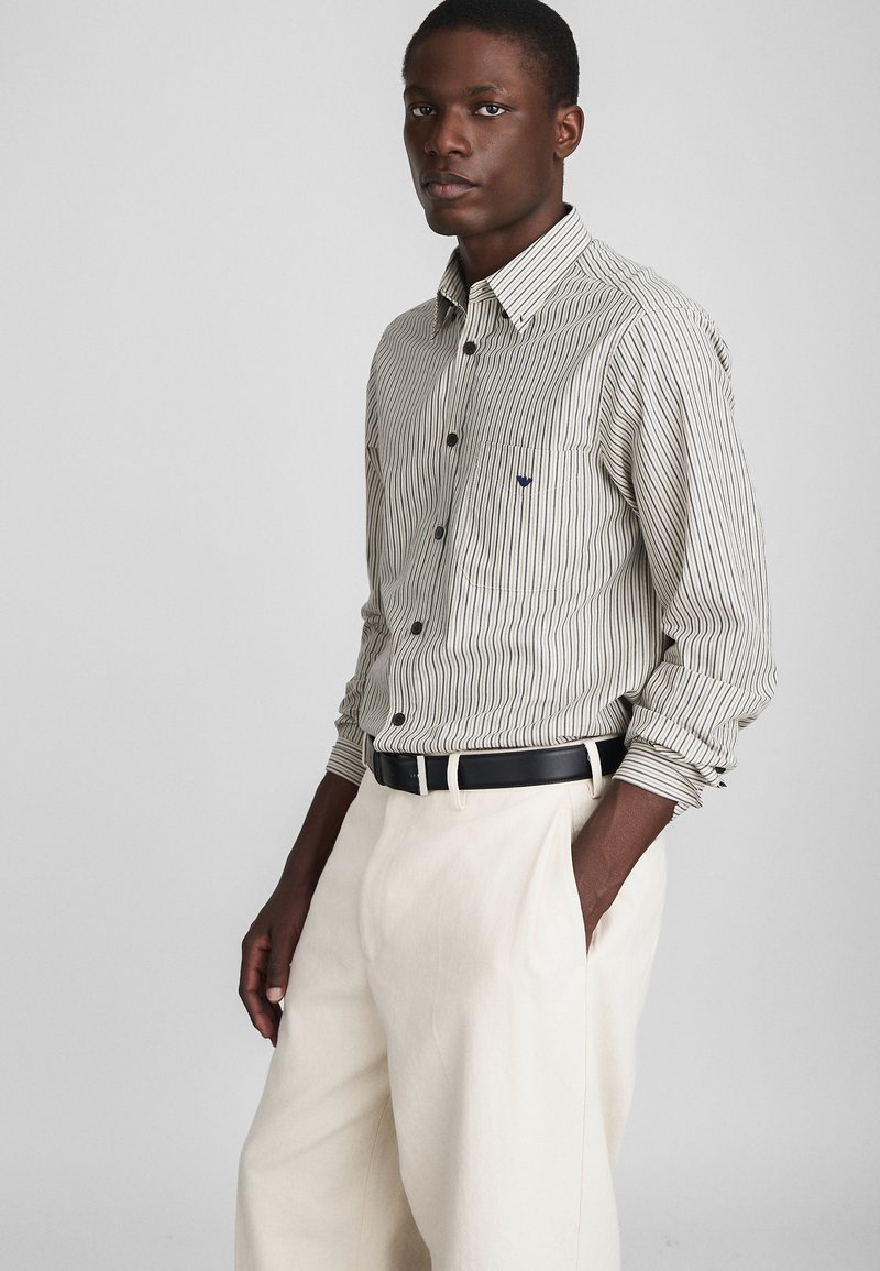 Svītraina krekla ar pogām krēmkrāsā un tumši zilā krāsā, ar kabatu un piegulošu piegriezumu, savienota ar krēmkrāsas biksēm un melnu jostu.