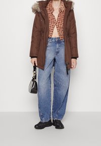 Hnědá zateplená parka s kapucí lemovanou kožešinou, košile s vzorem, volné modré džínové kalhoty, černé tenisky a malá černá kabelka.