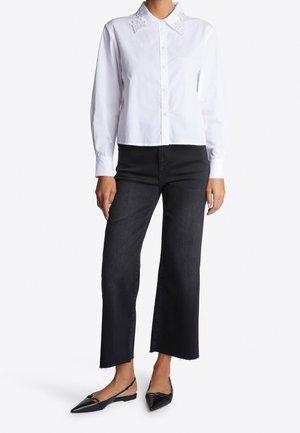 Vrouw die een wit overhemd met knopen draagt met kanten kraagdetails, zwarte cropped wijde jeans, en zwarte puntige slingback flats.