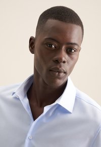 Camicia azzurra chiara con colletto strutturato, dotata di una fodera a fantasia in contrasto. Tessuto liscio e design aderente.