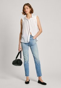 Hvid, ærmeløs skjorte med knaplukning og samlet frontdetalje, parret med lyseblå jeans, sort håndtaske og sorte, naglede flade sko.