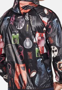 Veste noire présentant un collage de motifs graphiques colorés, incluant des outils, des vêtements et des accessoires. Légère, avec capuche et fermetures éclair.