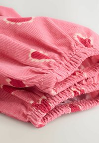 Rosa kordfloret tyg med röda hjärtmönster. Har elastiska samlingar upptill. Mjuk textur med synliga åsar.