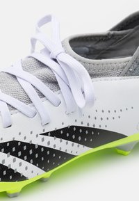 Scarpa da calcio bianca con tomaia in rete grigia e strisce diagonali nere. Presenta lacci bianchi, superficie testurizzata e suola verde neon.