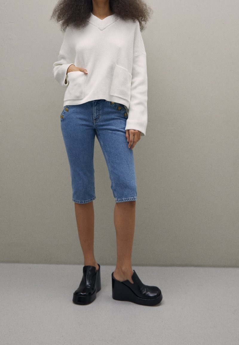 Pull blanc côtelé avec un col en V et une poche avant, associé à un short en denim bleu clair longueur genou avec des boutons en accent, et des chaussures noires à plateforme.