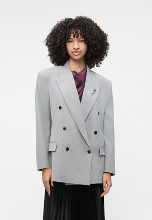 Grå dubbelknäppt blazer med svarta knappar, textil med struktur, breda slag och två framfickor, bärs över en mörklila topp.