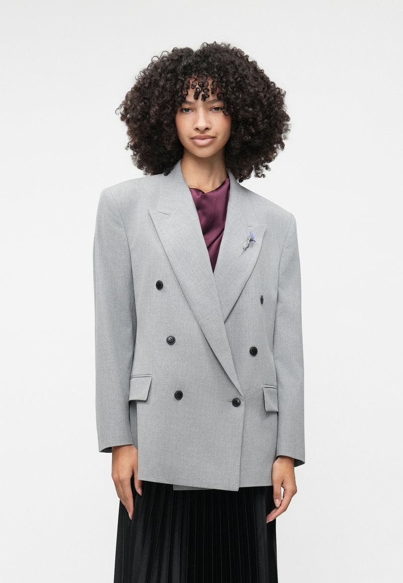 Harmaa kaksirivinen bleiseri mustilla napilla, teksturoitua kangasta, leveät kaulukset ja kaksi etutaskua, päällä tumman violetin värinen paita.