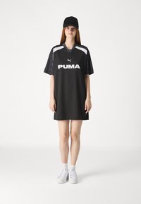 Vestido preto com mangas curtas, logótipo branco da PUMA, decote em V e detalhes pretos em padrão nas mangas. Combinado com ténis brancos e um boné preto.