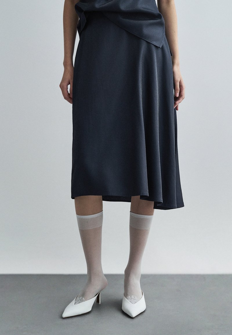 Mornarska midi krila s tekočim dizajnom, kombinirana s prosojnimi sivimi nogavicami in belimi čevlji na peto z oblikovanjem v obliki zaostrenega prsta, ki vsebujejo dekorativni srebrni dodatek.