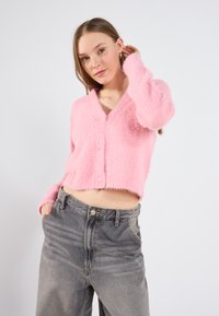 Mīksts rozā īsais kardigans ar garām piedurknēm, V veida kakla izgriezumu un pogu aizdari; kombinēts ar augsto jostasvīru pelēkiem džinsiem.