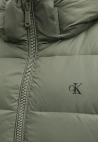Gros plan sur une doudoune verte avec une fermeture éclair visible et un logo Calvin Klein noir sur la poitrine.