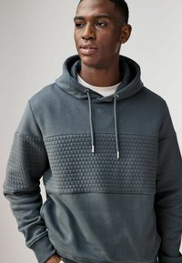 Grijze hoodie met een getextureerd honingraatachtig patroon aan de voorkant, met trekkoorden en een gladde afwerking op de mouwen en het onderste gedeelte.