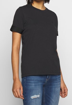 Vrouw draagt een effen zwart T-shirt met korte mouwen en blauwe jeans met een klein gescheurd plekje op het dijbeen.