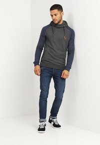 Sudadera con capucha de cuerpo gris y mangas azules, con cordón y un aspecto texturizado. Combinada con jeans de lavado oscuro y zapatillas.