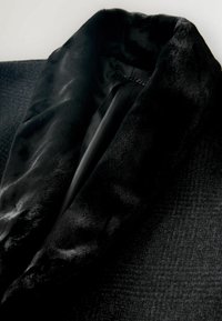 Čierny kabát s mäkkým umelým kožušinovým golierom, textúrovanou tkaninou s jemným vzorom a hladkou podšívkou.