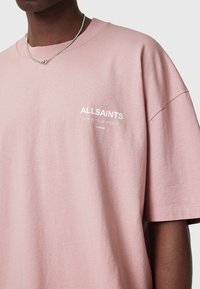 Camiseta rosa de algodón con un corte holgado, con un logotipo blanco que dice "ALLSAINTS" junto con texto adicional. Mangas cortas, cuello redondo.