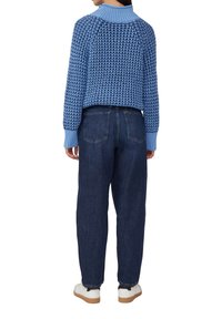 Persoon in een blauw gebreid sweater met patroon, donkere spijkerbroeken en witte sneakers, staande met de rug naar de camera op een witte achtergrond.