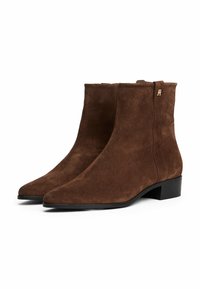 Stivali ankle in suede marrone con tacco nero basso, punta appuntita e piccolo dettaglio in oro vicino al bordo superiore.