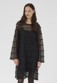 Melna kleita ar caurspīdīgu slāņveida audumu, piedāvājot ģeometrisku rakstu un frillēm gar piedurknēm un apakšmalu. Apaļš kakla izgriezums.