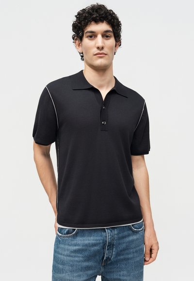 Νέος άνδρας με κατσαρά μαλλιά, φορώντας μαύρο κοντομάνικο πόλο με λευκά ράμματα, σε συνδυασμό με μπλε τζιν, στέκεται μπροστά σε ένα απλό φόντο.