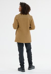 Braune Fleecejacke aus strukturiertem Stoff, verlängerte Länge und Seitentaschen. Kombiniert mit schwarzen Hosen und robusten, schwarzen Schuhen.