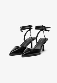 Chaussures à talons hauts en cuir noir avec un bout pointu et des brides de cheville. Le design présente un talon fin et une texture lisse avec des accents minimaux.