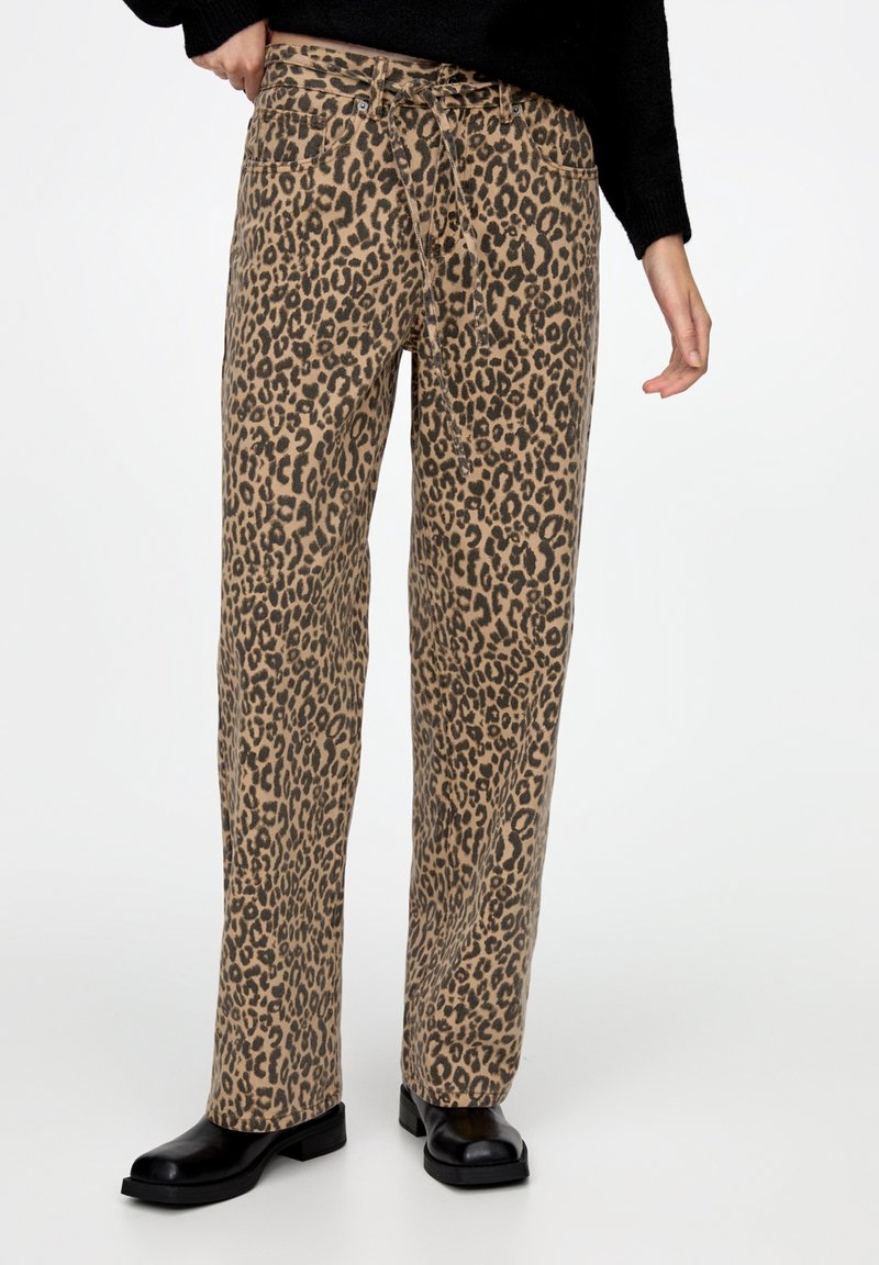 Pantalon imprimé léopard sur une base beige, avec une coupe décontractée, une taille à cordon de serrage et deux poches avant. Porté avec des chaussures plates noires.