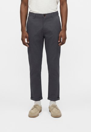 Homme portant un pantalon gris foncé à revers, des chaussettes blanches, des mocassins beiges et une chemise blanche à manches courtes, présenté de la taille aux pieds sur fond blanc.