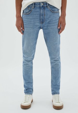 Jean slim bleu clair porté avec des baskets blanches sur une personne debout devant un fond uni.