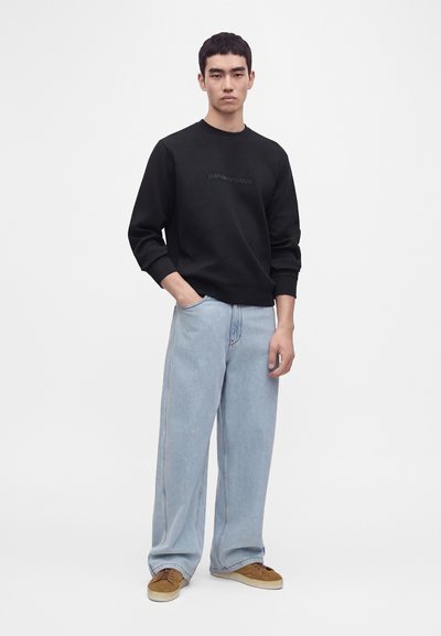 Jeune homme debout portant un sweat-shirt noir, un jean large bleu clair et des baskets marron sur un fond blanc uni.