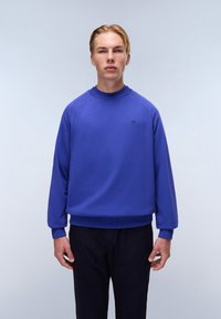Felpa blu con un collo alto, polsini e orlo a costine; presenta un piccolo logo sul petto. Abbinata a pantaloni scuri.