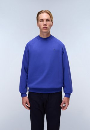 Blauwe sweatshirt met een hoge halslijn, geribde manchetten en onderrand; voorzien van een klein logo op de borst. Gepaard met donkere broeken.