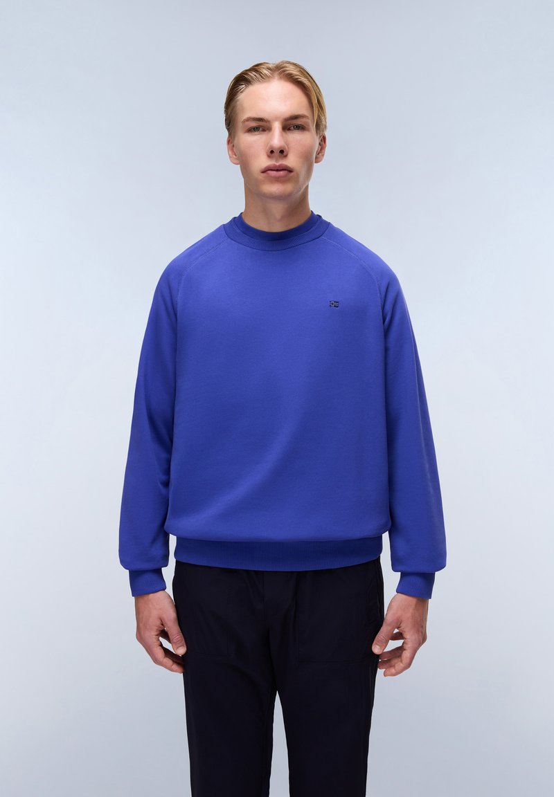 Felpa blu con un collo alto, polsini e orlo a costine; presenta un piccolo logo sul petto. Abbinata a pantaloni scuri.