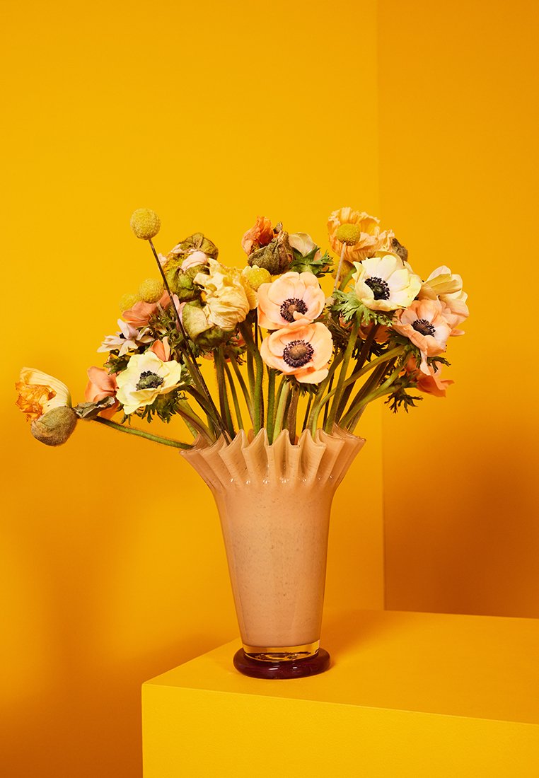 Pfirsich- und weiße Anemonenblumen mit grünen Blättern und gelben Billy-Bällen, angeordnet in einer gerillten Pfirsichvase auf einer gelben Oberfläche und einem gelben Hintergrund.